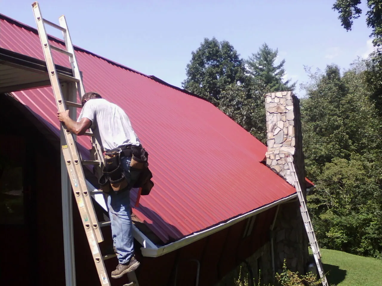 Roofer on a ladder beside a red metal roof and stone chimney — Soffit & Fascia Repair by Roofing Brothers in Ashaway